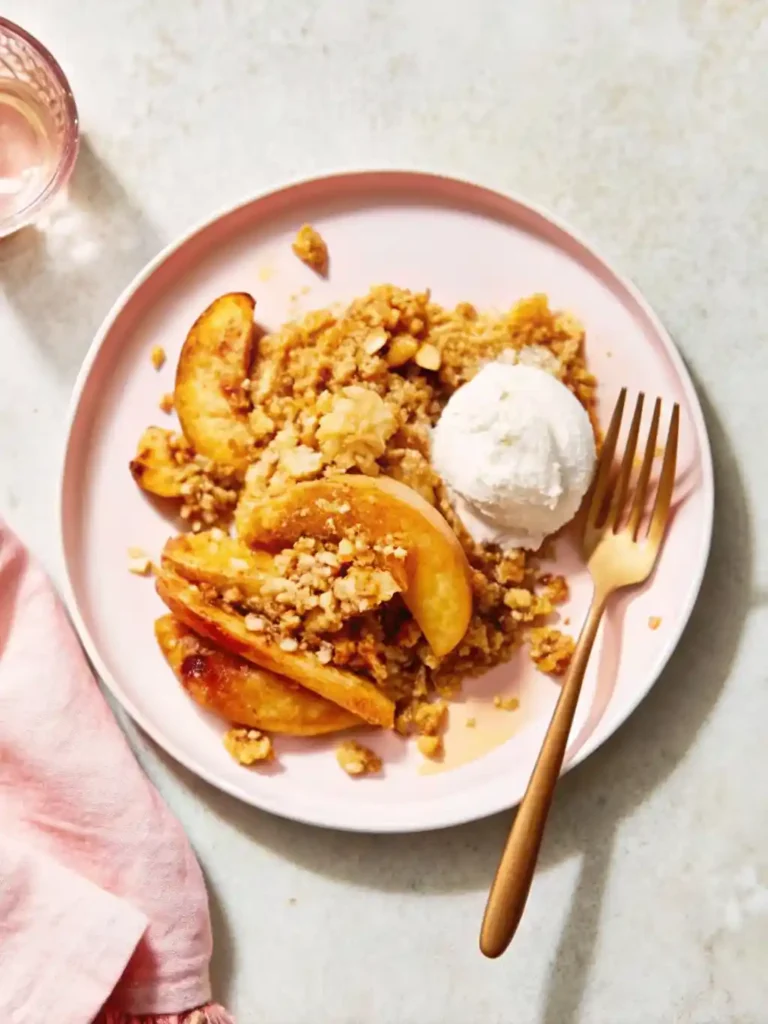 A serving of warm peach crisp topped with a scoop of vanilla ice cream on a pink plate—an irresistible peach recipes dessert perfect for summer.