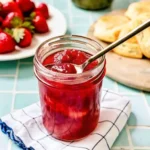 Jar of homemade strawberry rhubarb jam recipe with spoon, served alongside fresh strawberries and fluffy biscuits.