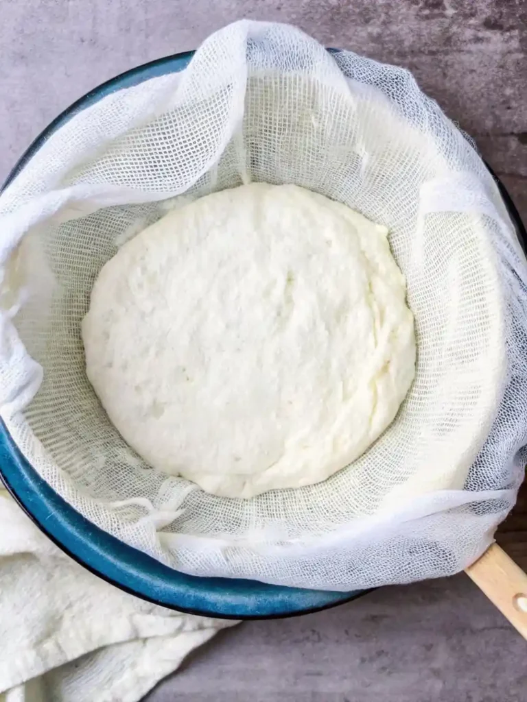 Homemade Greek yogurt being strained through cheesecloth in a blue colander to achieve thick, creamy texture.