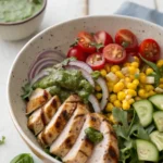 Close-up of grilled chicken salad with corn, cucumber, red onion, and basil dressing in a ceramic bowl on white table