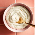 Smooth and thick homemade Greek yogurt in a pink bowl with a golden spoon on a pink tile background.