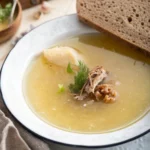 Homemade Jewish chicken soup recipe with dill and shredded chicken, served alongside a slice of brown bread on a white plate with a blue rim.