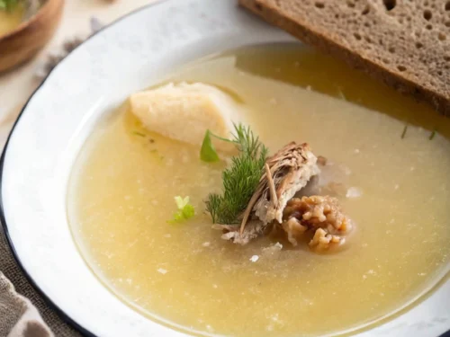 Homemade Jewish chicken soup recipe with dill and shredded chicken, served alongside a slice of brown bread on a white plate with a blue rim.