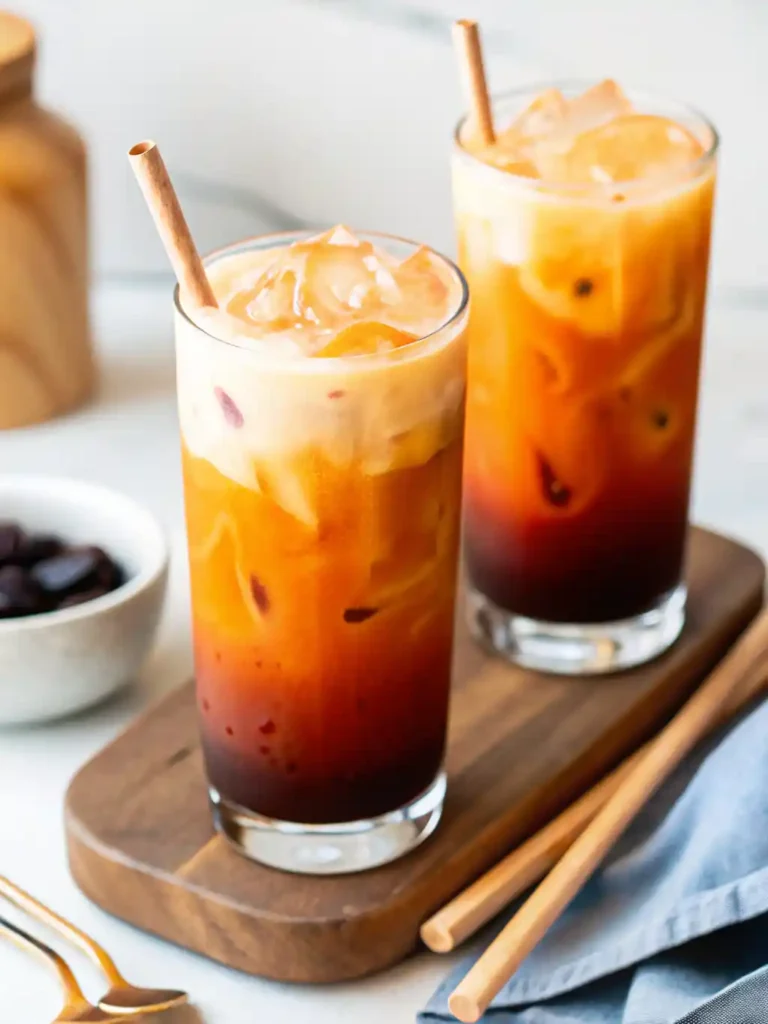 Two tall glasses of Thai iced tea with ice and creamy milk swirls, served with wooden straws on a wooden board.
