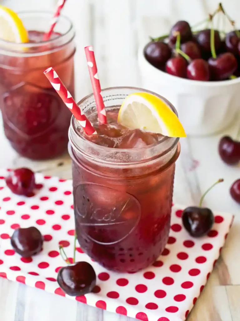 Homemade cherry iced tea served in mason jars with lemon slices and red straws, surrounded by fresh cherries, perfect for summer recipe ideas.